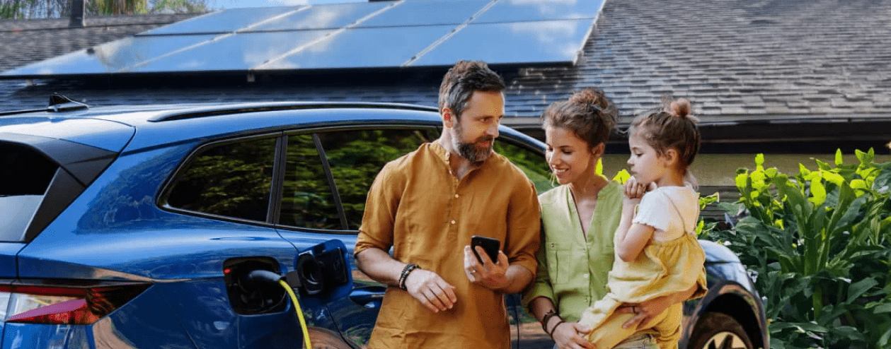 A family stands next to a pluggedin electric vehicle while a home with rooftop solar panels is in th